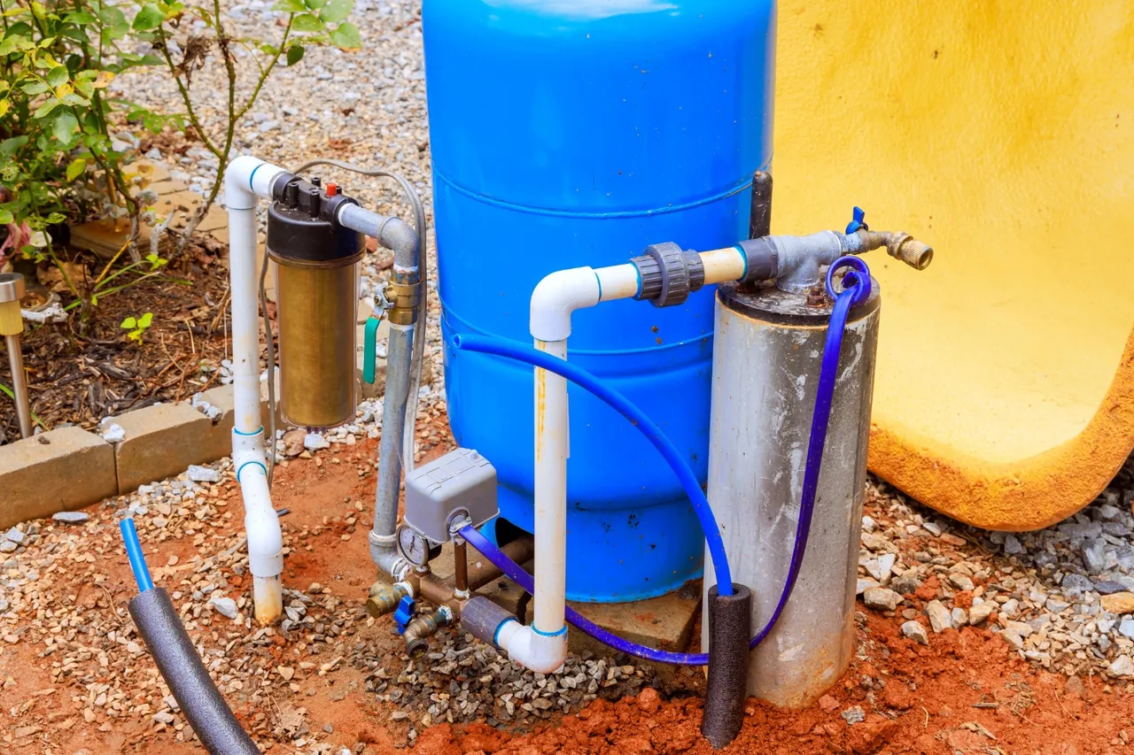 Sistema de pressurização de poço instalado em quintal, com tanque azul, filtro e tubulação de PVC