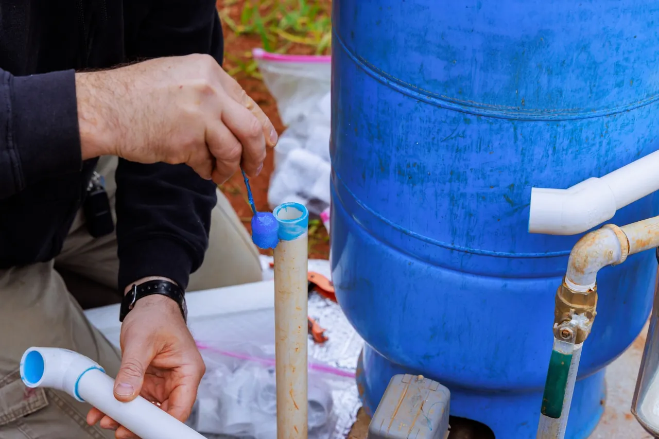 Mão de técnico com luva colocando tampa em tubo de PVC próximo ao tanque azul, manutenção de bomba de poço no RJ